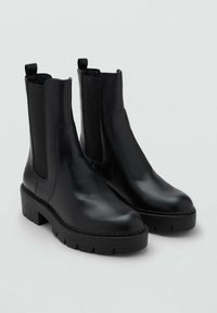 Botine Chelsea din piele neagră cu tălpi groase, panouri elastice pe laturi și mânere de prindere pe un fundal gri deschis.