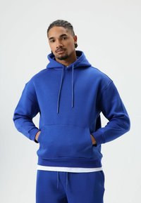 Blue hoodie with a drawstring hood, front pocket, and ribbed cuffs. Made from soft material, features a relaxed fit. Paired with blue joggers.