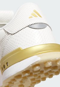White golf shoe with textured, reptile-like upper, gold accents, gray detail, and a patterned rubber outsole featuring small circular grips.