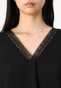 Schwarze Bluse mit V-Ausschnitt, verziert mit filigranem schwarzen Spitzenbesatz und floralen Mustern. Weiche Textur mit leichtem Glanz. Keine weiteren Details sichtbar.