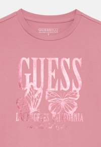 Rožinė organinės medvilnės marškinėlių su ryškiu "GUESS" logotipu blizgia balta spalva, su drugelių grafika ir tekstu, nurodančiu "Los Andželas, Kalifornija."