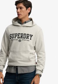 Sudadera gris con capucha y logo negro "SUPERDRY", bolsillo canguro frontal, llevada sobre una camisa azul a cuadros, combinada con jeans oscuros.