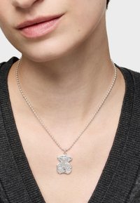 Colgante en forma de oso plateado sobre un collar de cuentas delgadas, reposando sobre un suéter gris oscuro. El colgante tiene un acabado de superficie texturizada.