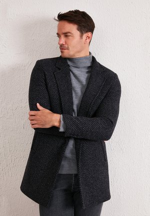 Dunkel strukturierter Wollmantel mit einer einzigen Knopfverschluss, Reverskragen und tailliertem Schnitt, getragen über einem hellgrauen Rollkragenpullover.