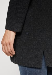Mørkegrå strikket cardigan med ribbede manchetter og et lille mærke, samt en let længere bagkant til lagdeling.