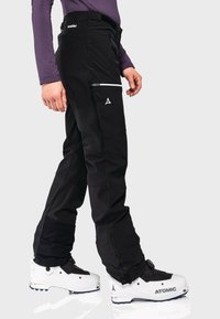 Schwarze Skihosen mit einer Reißverschlusstasche, aus einem strapazierfähigen Stoff, kombiniert mit weißen Skischuhen mit schwarzem Akzent.