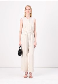 Combinaison en lin beige avec boutons et ceinture à nouer, dotée de jambes larges. Associée à un petit sac à main noir et des sandales nude.