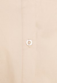 Camicia beige con bottoni a quattro fori di colore bianco, cuciture visibili e un tessuto liscio e strutturato.