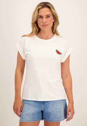 Vrouw met blond haar die gouden creolen draagt, wit T-shirt met watermeloenpatch en blauwe denimshorts tegen een effen achtergrond.