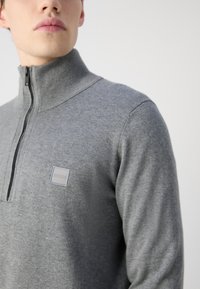 Pilkas zip-up megztinis, pagamintas iš minkštos medžiagos, su raštuotu apykakle, siaura siluetu ir mažuoju kvadratiniu logotipu ant krūtinės.