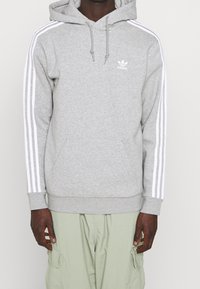 Persona vestida con una sudadera gris de Adidas con rayas blancas en las mangas y pantalones cargo verdes claros, de pie contra un fondo sencillo.