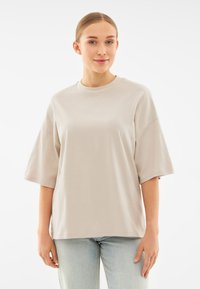 Tricou bej cu maneci scurte, confecționat dintr-un material moale, având o gât rotund, o croială lejeră și umeri ușor lăsați.