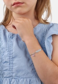Bracciale in argento con un'etichetta rettangolare e un piccolo charm rosa. La persona che lo indossa ha la pelle chiara ed è vestita con una blusa azzurro chiaro con volant.