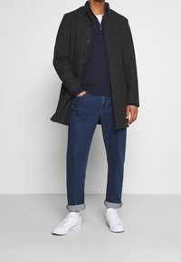 Cappotto in misto lana grigio con collo alto, indossato sopra un maglione blu navy, abbinato a jeans blu e sneakers bianche. Look casual e strutturato.