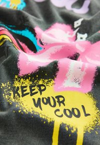 Čierna tkanina s živým graffiti dizajnom. Obsahuje ružové, žlté a modré škvrny. Text znie "Udržuj si chlad" v tučnom písmene.