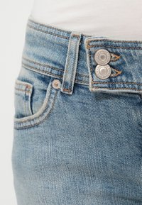 Jean en denim bleu présentant une coupe fuselée avec une texture délavée, deux boutons argentés et des coutures contrastées orange le long des coutures.