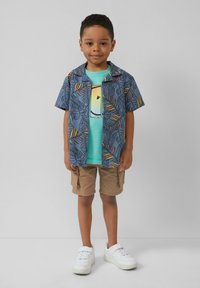 Korte mouwen blauwe shirt met kleurrijk bladerenpatroon over een turquoise t-shirt, beige cargoshorts en witte sneakers.