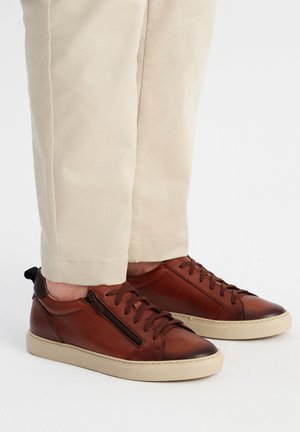 Brązowe skórzane trampki o teksturowanym wykończeniu, z bocznym zamkiem błyskawicznym, sznurowanym przodem i kremową podeszwą. Noszone z jasnobeżowymi spodniami.