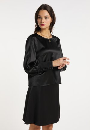 Femme portant un chemisier en satin noir à manches longues avec poignets boutonnés et une jupe noire assortie, accessoirisée de bijoux en or.