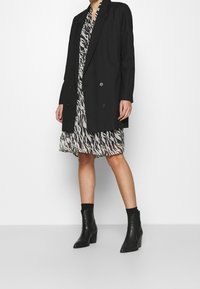 Černý oversized blazer s dvojitými knoflíky přes vzorované, monochromatické šaty. Modelka nosí černé kotníkové boty s mírným podpatkem.