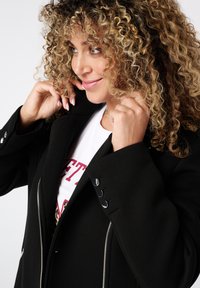 Zwarte blazer met een gestructureerd ontwerp, voorzien van zilveren rits- en knoopaccenten. Draagt over een wit T-shirt met vetrode tekst. Krullend haar zichtbaar.