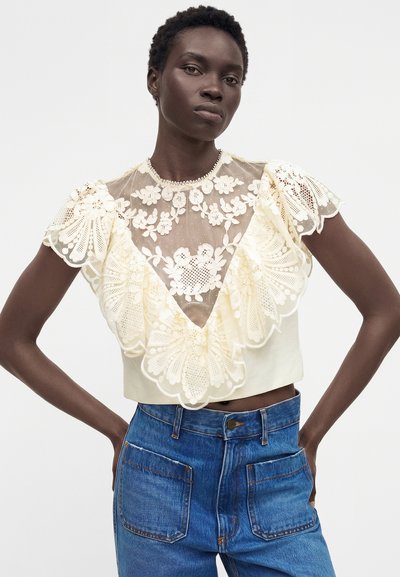 Γυναίκα που φοράει μια διαφανή εκρού φλοράλ δαντελένια μπλούζα με βολάν στα μανίκια, συνδυασμένη με ψηλόμεσα μπλε τζιν, όρθια με τα χέρια στη μέση.