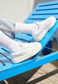 Baskets blanches avec un dessus texturé et une semelle en caoutchouc, présentant des découpes subtiles et un accent rose clair au niveau du talon, associées à des chaussettes blanches.