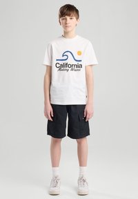 Valkoinen puuvillapaita, jossa on sininen aalto- ja auringografiikka, yhdistetty mustiin cargo-shortsihin, valkoisiin sukkiin ja valkoisiin lenkkareihin. Yksinkertainen ja rento muotoilu.