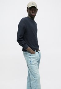 Tamsiai mėlynas megztinis, šviesiai mėlynos spalvos džinsai, smėlio spalvos kepurė. Modelis stovi šonu, rankos kišenėse, demonstruodamas laisvalaikio aprangą.