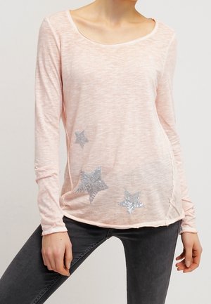 Frau trägt ein hellrosa Langarmshirt mit silbernen Sternmotiven auf der Vorderseite, kombiniert mit dunkelgrauen Hosen.