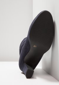 Bottine à talon moyen en daim noir montrant la semelle texturée et la taille 37, inclinée contre un fond de sol et mur blanc.