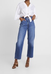 Valkoinen napillinen paita, solmittu vyötärölle, vaaleansiniset korkeavyötäröiset farkut ja ruskeat leopardikuosiset leveäkorkoiset sandaalit.