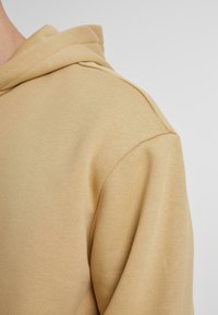 Gros plan d'un sweat à capuche beige montrant la couture de l'épaule et une partie de la capuche sur un fond uni.