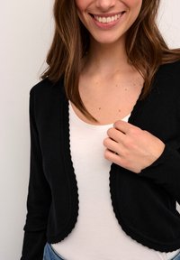 Černý zkrácený kardigan s vlnitými okraji, z texturované pleteniny, nošený přes bílý top. Modelčina ruka se dotýká přední části kardiganu.