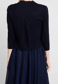 Marinblå stickad kort cardigan med trekvartsärmar, som har en slät yta och knäppningsdetaljer på ärmarna.