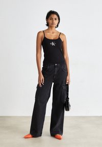 Black tank top with thin straps and logo, paired with black wide-leg cargo pants featuring side pockets. Bright orange pointed shoes.