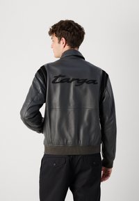 Zwarte leren jack met geribde onderkant en manchetten; heeft suede schouderaccenten en "targa" borduurwerk op de achterkant. Glad van textuur.