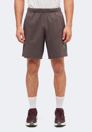 Homme portant un short gris foncé au niveau des genoux, des chaussettes blanches et des chaussures de course bordeaux, debout avec les mains détendues le long du corps.