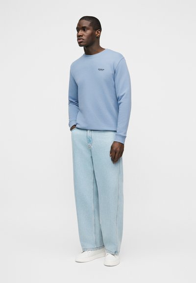 Άνδρας που φοράει γαλάζιο φούτερ, φαρδιά τζιν παντελόνια ανοιχτού χρώματος, λευκά αθλητικά παπούτσια, στέκεται με το ένα χέρι στην τσέπη, κοιτώντας προς τα αριστερά.