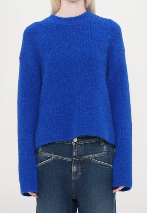 Kobieta w jasnoniebieskim puszystym swetrze z długimi rękawami, w połączeniu z ciemnoniebieskimi jeansami z wysokim stanem.