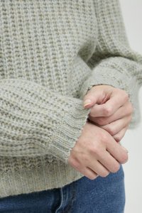 Hellgrauer Strickpullover mit einem strukturierten Muster, ausgestattet mit gerippten Bündchen und einer weichen, flauschigen Oberfläche. Hände greifen nach dem Ärmel.