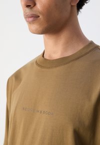 Barna pamut póló kerek nyakkal és rövid ujjal. Elöl egy egyszerű, visszafogott betűtípussal készült szürke "SCOTCH&SODA" logó található.