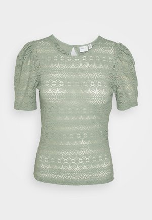 Blusa de encaje verde claro con mangas cortas, con estampados florales y una pequeña abertura en forma de ojo de cerradura en la parte trasera del cuello.