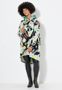 Robe à capuche noire avec des imprimés graffiti multicolores. Manches longues, ourlet plus long à l'arrière, associée à des leggings noirs brillants et des bottes cloutées.