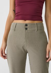 Vrouw in hoog getailleerde beige en zwart geblokte broek met twee zwarte knopen, gecombineerd met een kort bordeauxrood topje dat een vlinder tatoeage toont.