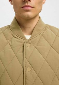 Kurtka bomber z pikowanej beżowej tkaniny z prążkowanym kołnierzem, wyposażona w dwa zatrzaski oraz gładki, teksturowany materiał.