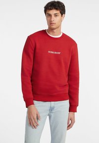 Rød hettegenser laget av bomull, med rund hals og ribbestrikkede mansjetter. Fronten viser "GUESS JEANS" -logoen i hvit brodering. Kombinert med lyseblå jeans.