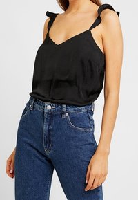Top nero senza maniche con scollatura a V, caratterizzato da spalline arricciate. Abbinato a jeans in denim blu a vita alta. Texture del tessuto liscia.