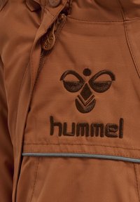 Bruine jack met een rits, gemaakt van een textuurstof. Geborduurd logo in donkerbruin op de borst en grijze accenten aan de onderkant.