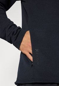 Dunkelblauses Houdini-Shirt aus strukturiertem Stoff, mit langen Ärmeln und einer Reißverschlusstasche. Mit gesticktem Logo auf der linken Seite.